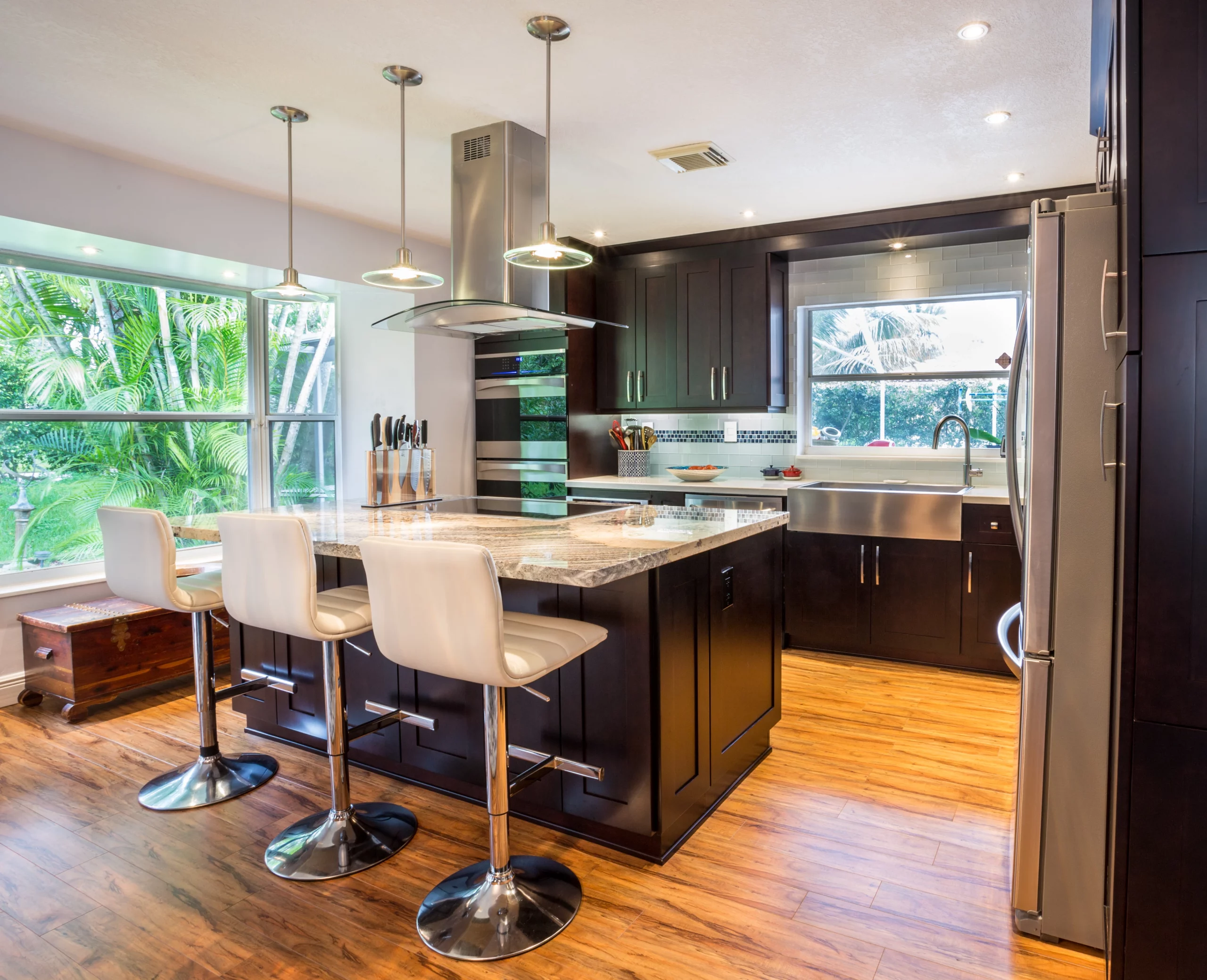 Modern kitchen island with dark cabinets, granite counters, and white bar stools overlooking lush greenery.