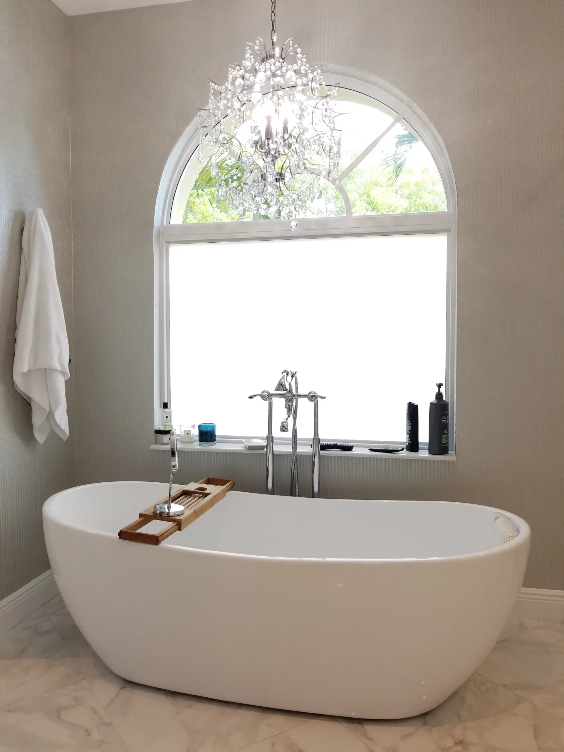 Luxury freestanding tub in an elegant bathroom remodel featuring a crystal chandelier and arched window.