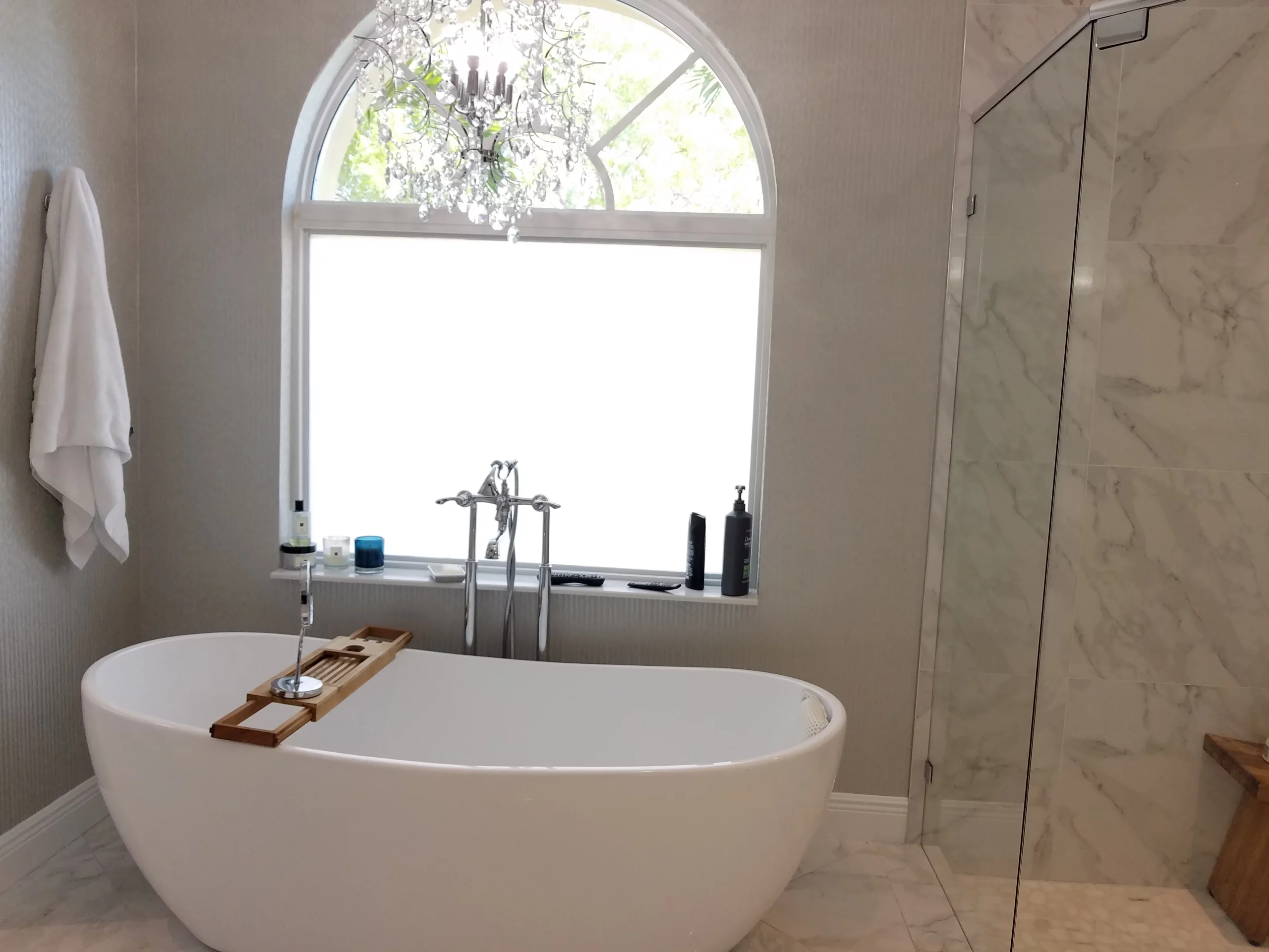 Luxury freestanding tub beneath an arched window with a crystal chandelier in an elegant bathroom remodeling design.