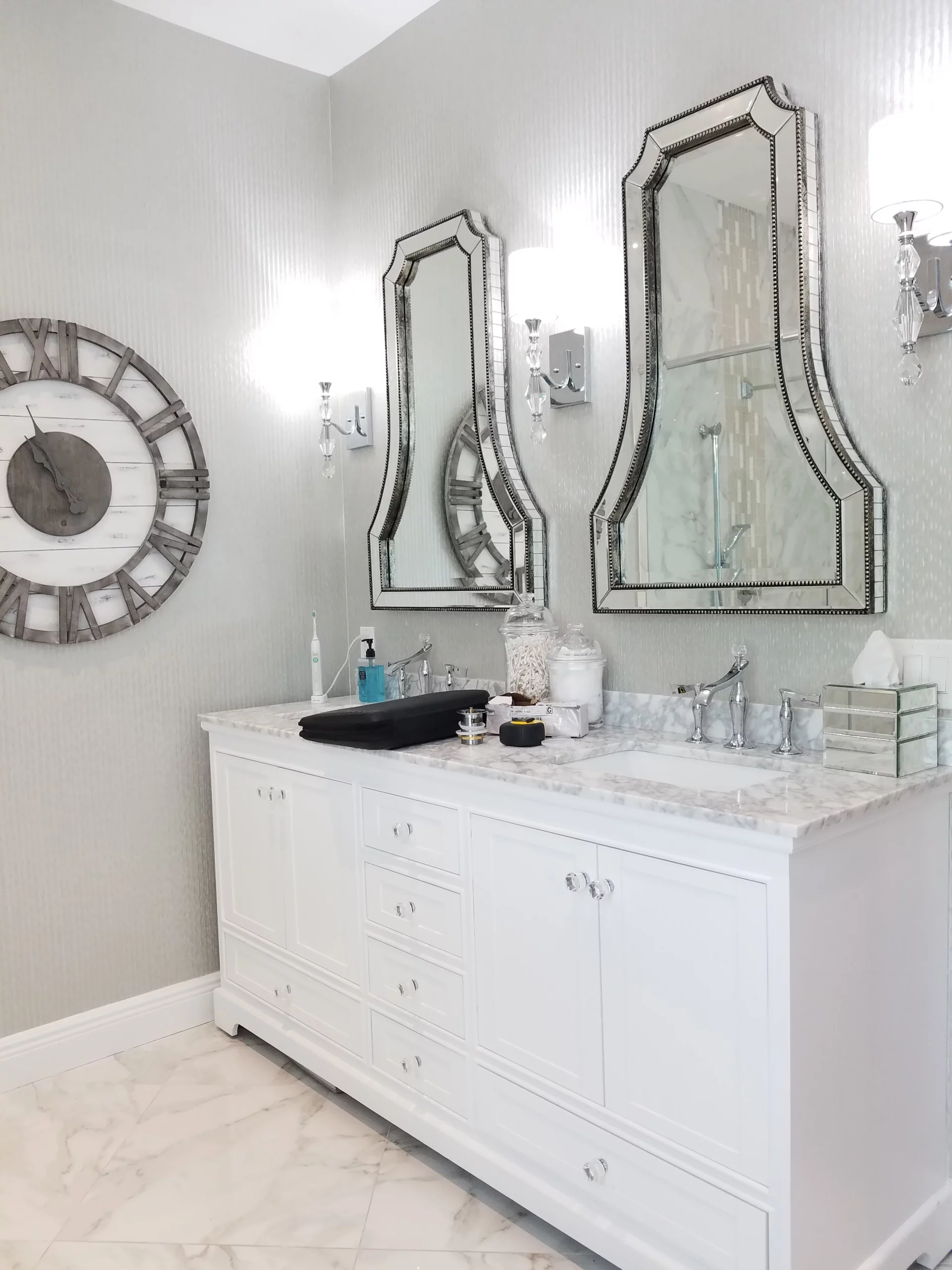 White double vanity with marble top, mirrored sconces, and large Roman numeral clock in a bathroom remodeling.