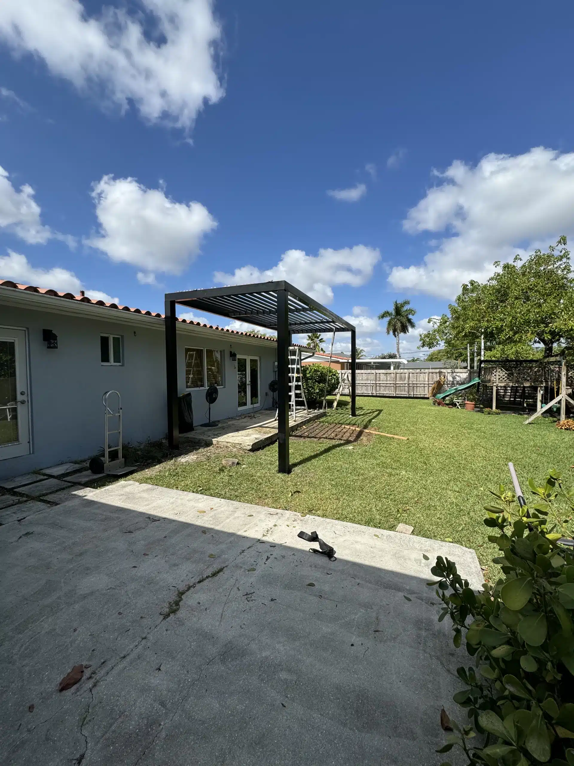 Black aluminum solid-roof pergola providing full sun and rain protection over a concrete patio next to a residential swimming pool.