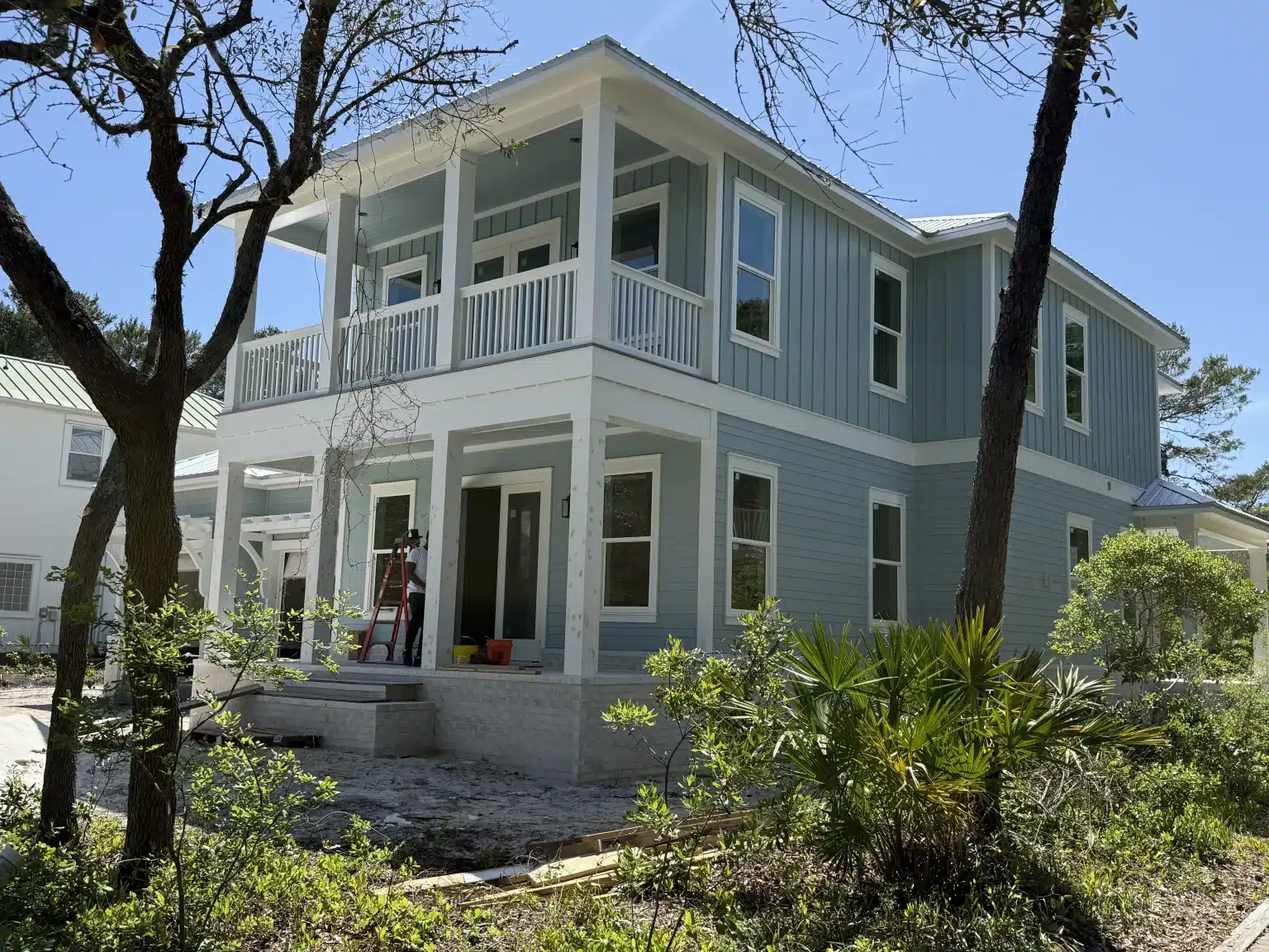 Modern two-story coastal style home during new home construction, featuring white wrap-around porches and light siding surrounded by trees.
